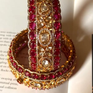 Puede incluir: Dos brazaletes dorados con diseños intrincados. Los brazaletes están adornados con piedras preciosas rojas de forma ovalada y piedras claras, cuadradas y con forma de flor. Las joyas se exhiben en un libro.