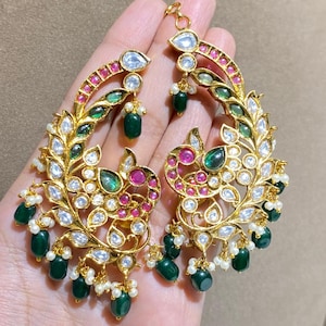 Puede incluir: Pendientes dorados ornamentados con un diseño de hojas. Los pendientes presentan piedras preciosas transparentes, verde esmeralda y color rubí, junto con pequeñas perlas blancas. Los pendientes se sostienen en una mano.