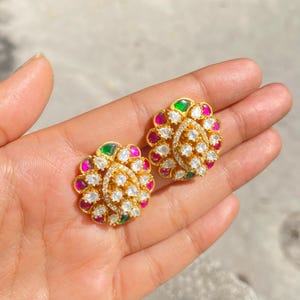 May include: A pair of gold-colored stud earrings with a floral design. Each earring features a central cluster of white stones surrounded by alternating pink, green, and white stones. The earrings are held in a hand.