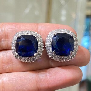 May include: A pair of square-cut, dark blue gemstone stud earrings. Each earring is surrounded by a halo of small, clear stones set in a silver-colored metal. The earrings are displayed on a light-colored background.