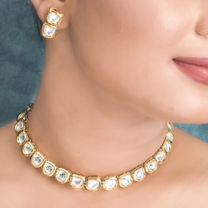 May include: A gold necklace with a row of clear, faceted stones. The necklace is on a model's neck. The model is wearing matching earrings with clear stones.