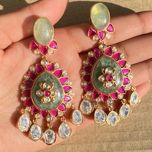 Puede incluir: Un par de pendientes colgantes con piedras verdes y rosas. Los pendientes tienen una piedra verde en forma de lágrima rodeada de piedras rosas y una piedra blanca más pequeña en el centro. Los pendientes están adornados con piedras transparentes.