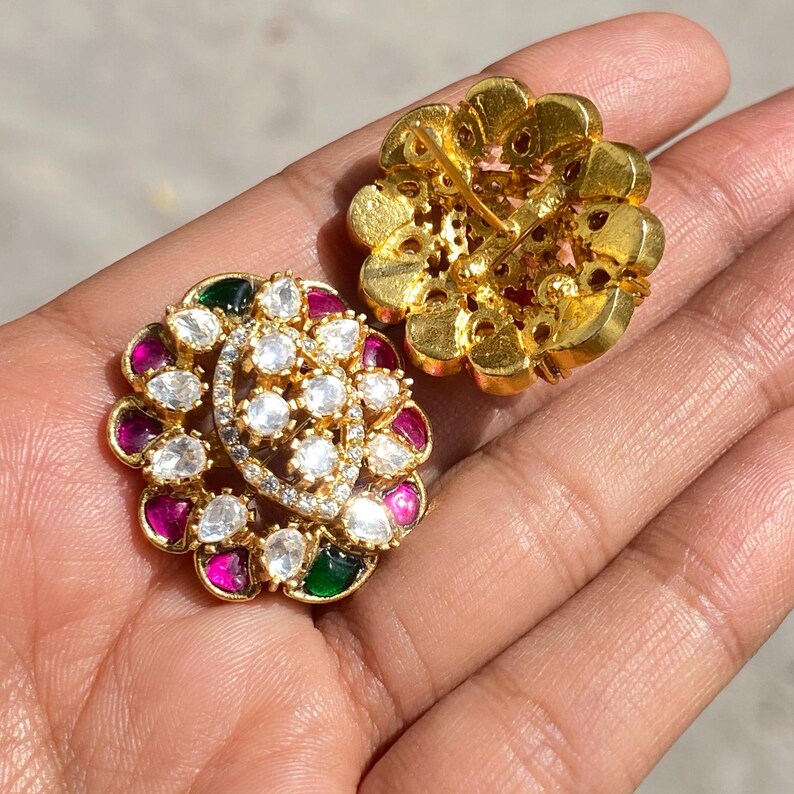 May include: Two gold-toned flower-shaped earrings. Each earring features a central cluster of clear, round stones surrounded by smaller, colorful stones in shades of red, green, and clear. The earrings are designed with a floral motif.