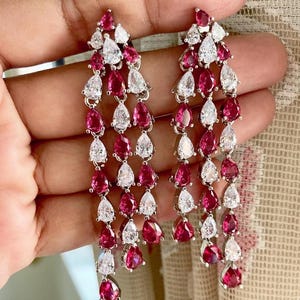 Pode incluir: Um par de brincos longos e pendentes com uma cascata de pedras vermelhas e transparentes em forma de lágrima. Os brincos são prateados e apresentam um grupo de pedras na parte superior.