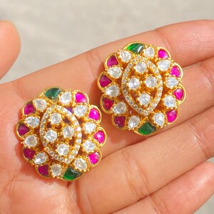 May include: A pair of gold-colored stud earrings. Each earring features a floral design with a central oval shape encrusted with small white stones. The petals are adorned with alternating pink, green, and white stones, creating a colorful and intricate pattern. The earrings are set against a neutral background. Green Kundan Earrings Emerald Polki Earrings Jadau Kundan Jewelry Navratan Earrings Indian Stud Earrings Indian Stud Kundan Kewelry