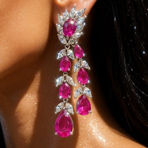 May include: Elegant drop earrings featuring vibrant pink teardrop-shaped gemstones. The earrings have a cluster of white gemstones at the top and are set in a silver metal. The design is a cascade of pink gems and white accents.