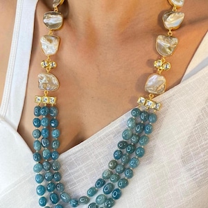 May include: A multi-strand necklace with blue gemstone beads and gold accents. The necklace features a center pendant with a large, white, irregular-shaped stone.