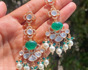 May include: Gold-toned chandelier earrings with emerald green and clear gemstone accents. The earrings feature a floral design with dangling white pearls and small green beads. The earrings are held in a hand.