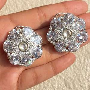 May include: Two flower-shaped stud earrings. Each earring features a central, clear, round stone surrounded by smaller, clear stones. The outer petals are formed by larger, clear, faceted stones, creating a sparkling, floral design. The earrings are set on silver posts.