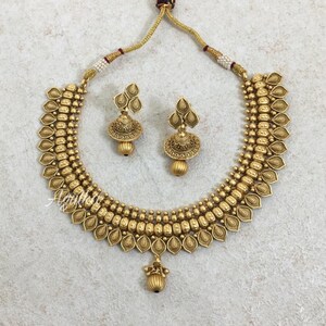May include: A gold-toned necklace and earring set. The necklace features a detailed design with leaf-shaped elements and a central pendant. The matching earrings have a similar design. The jewelry set is displayed on a light-colored surface.