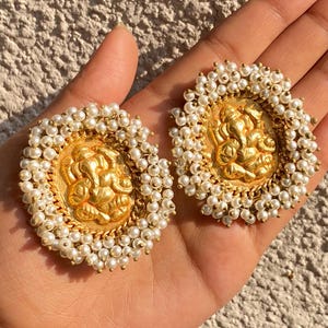 Puede incluir: Un par de pendientes redondos de tono dorado con una figura central en relieve de Ganesha rodeada por un borde denso de pequeñas cuentas blancas esféricas. El diseño circular tiene un acabado metálico texturizado.