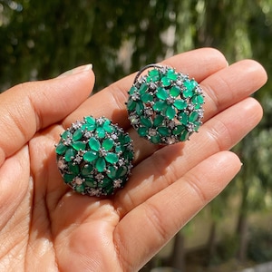 May include: A pair of dome-shaped stud earrings featuring clusters of marquise-cut green stones arranged in flower patterns, accented with small clear round stones set in a silver-toned metal base.