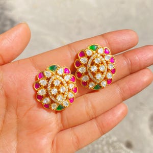 May include: A pair of gold-toned stud earrings with a floral design. Each earring features a central oval of small, clear stones surrounded by alternating pink, white, and green stones. The earrings are held in a hand.