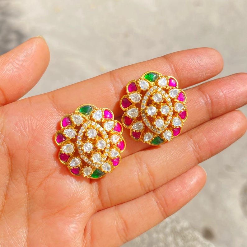 May include: A pair of gold-toned stud earrings with a floral design. Each earring features a central oval of small, clear stones surrounded by alternating pink, white, and green stones. The earrings are held in a hand.