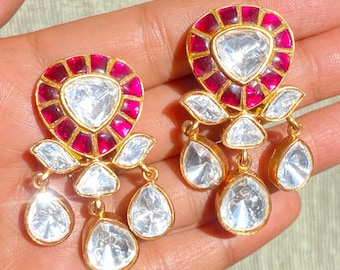 May include: A pair of gold-toned earrings with a central clear, faceted stone surrounded by a ring of red stones. The earrings have three dangling clear, faceted teardrop-shaped stones.