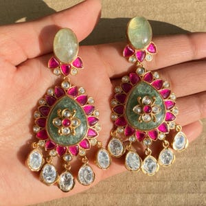 May include: A pair of dangle earrings with a green stone center surrounded by pink and white stones. The earrings have a gold finish.