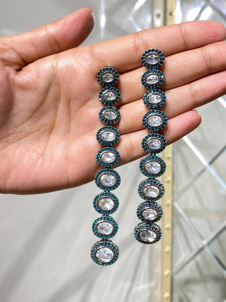 Pu&ograve; includere: Orecchini pendenti lunghi con una serie di pietre ovali trasparenti circondate da accenti color teal. Gli orecchini sono progettati con una disposizione lineare di pietre, creando un look sofisticato ed elegante. Un gioiello.