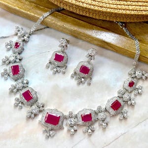 May include: A silver-coloured jewellery set comprising a necklace and matching earrings. The necklace and earrings feature large, square, ruby-red gemstones surrounded by smaller, clear stones. The set is displayed on a light-coloured surface.