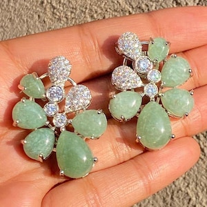 Può includere: Un paio di orecchini color argento con un design floreale. Ogni orecchino presenta pietre a goccia verde chiaro e cristalli trasparenti. Gli orecchini sono su uno sfondo neutro. Le pietre misurano circa 1 cm.