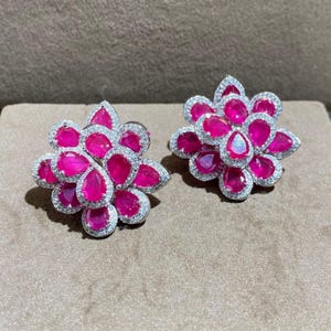 May include: A pair of floral-shaped stud earrings featuring vibrant pink pear-cut stones arranged in a cluster pattern. Each individual pink stone is surrounded by a delicate border of small, clear white pavé-set stones, creating a bright, sparkling contrast against the deep pink colour of the central gems.