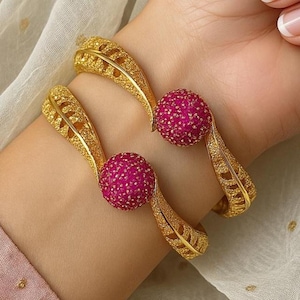 Puede incluir: Dos brazaletes dorados con un diseño de hojas y una esfera central magenta texturizada. Los brazaletes se llevan en una muñeca, mostrando los intrincados detalles de la joyería.