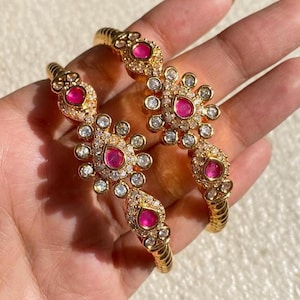 May include: Two gold bangles with floral designs. Each bangle features ruby-coloured gemstones surrounded by smaller, clear stones. The bangles have a textured, ridged design along the band.