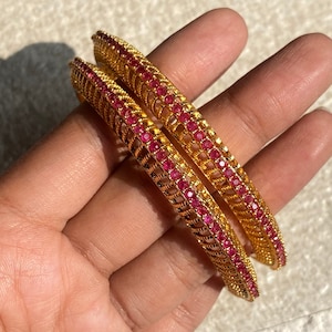 May include: Two gold-colored bangles with a repeating pattern of small, round, red gemstones. The bangles have an openwork design and are held in a hand.