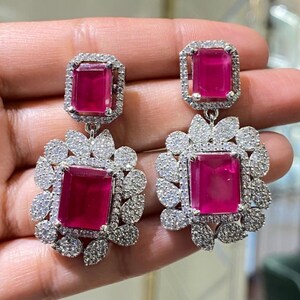 Puede incluir: Un par de pendientes colgantes plateados. Cada pendiente presenta una gran piedra rectangular de color magenta rodeada de pequeños cristales transparentes. La piedra inferior está engastada en un racimo floral de cristales.