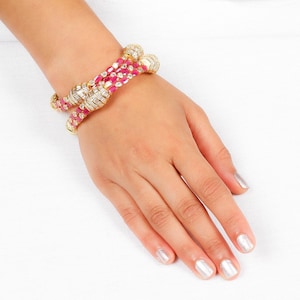 May include: A gold bangle bracelet featuring a central row of oval-cut pink gemstones flanked by two rows of clear baguette-cut stones. The design includes decorative gold sections set with clear stones at the ends of the bangle structure.