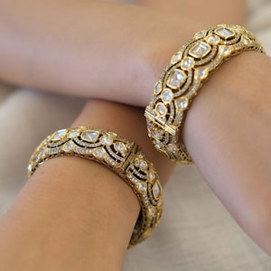 Pode incluir: Um par de pulseiras articuladas em tom dourado com um design intrincado e vazado. Cada pulseira é adornada com inúmeras pedras transparentes facetadas num padrão repetitivo, acentuadas por pequenas pedras pavé nas bordas metálicas.