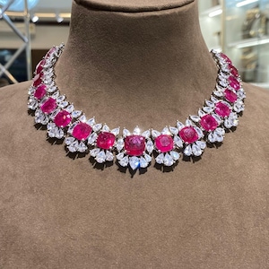 May include: A dazzling necklace featuring large, square-cut, ruby-red gemstones, interspersed with clusters of smaller, clear, white stones. The necklace is displayed on a brown mannequin head, showcasing its intricate design and vibrant colors.