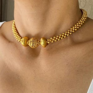 May include: A gold-colored necklace with a beaded design. The necklace features three larger, textured beads in the center, flanked by smaller, round beads. The necklace is worn around the neck.