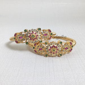 May include: Two gold-tone bangles featuring intricate floral-inspired metalwork, inlaid with small clear crystals, round pink stones, and tiny green accents. The central design is ornate and symmetrical, with the script text Agulka visible.