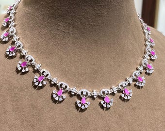 May include: A silver-toned necklace with a series of fan-shaped pendants, each featuring a vibrant pink gemstone at its center. The necklace is adorned with numerous clear, sparkling stones, creating a dazzling effect. The design is symmetrical and elegant.