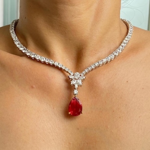 May include: A diamond necklace with a teardrop ruby pendant. The necklace is set with round-cut diamonds in a delicate, curved design. A large, vibrant red ruby pendant hangs from the centre, providing a striking contrast to the clear diamonds.