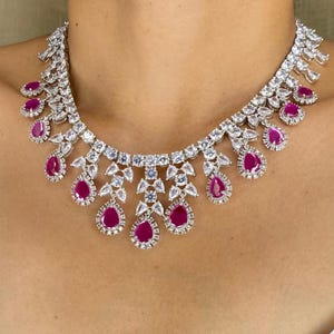 Puede incluir: Un collar deslumbrante con una serie de piedras claras y brillantes y gemas de color rojo rub&iacute; en forma de l&aacute;grima. El collar est&aacute; dise&ntilde;ado para colocarse alrededor del cuello, con las gemas de rub&iacute; cayendo.
