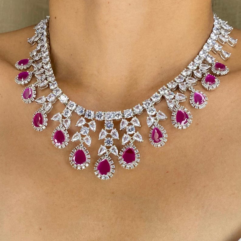 Puede incluir: Un collar deslumbrante con una serie de piedras claras y brillantes y gemas de color rojo rub&iacute; en forma de l&aacute;grima. El collar est&aacute; dise&ntilde;ado para colocarse alrededor del cuello, con las gemas de rub&iacute; cayendo.