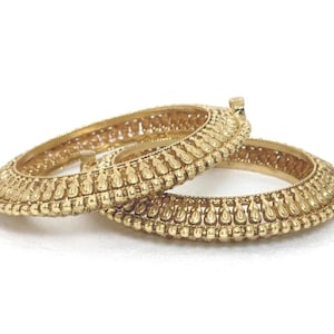 May include: Two gold-toned bangles featuring a raised, repeating teardrop pattern along the outer edge. Each bangle has a textured, beaded border and a small, cylindrical protrusion extending from the side of the circular metal frame.