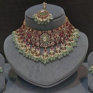 Puede incluir: Un conjunto de collar de novia tradicional indio con un collar dorado, pendientes y una diadema. El collar está adornado con piedras preciosas rojas y verdes, perlas y diseños florales intrincados. Los pendientes y la diadema presentan diseños y adornos a juego.