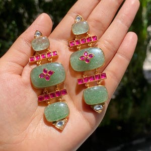 Puede incluir: Un par de pendientes colgantes dorados. Cada pendiente presenta tres piedras rectangulares de color verde claro, conectadas por barras doradas con pequeñas piedras rosas. Los pendientes tienen un diseño floral en el centro y una piedra transparente en la parte superior e inferior.