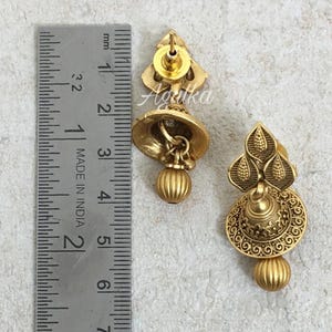 May include: Gold-toned earrings with intricate detailing. The earrings feature a floral design at the top, a bell-shaped center, and a spherical bead at the bottom. The earrings are approximately 1 inch in length. The text "Agulka" is visible on the earrings.