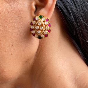 May include: Gold-toned stud earrings with a floral design. The earrings feature a central cluster of clear, sparkling stones surrounded by alternating pink, green, and clear stones. The earrings are set against the skin.