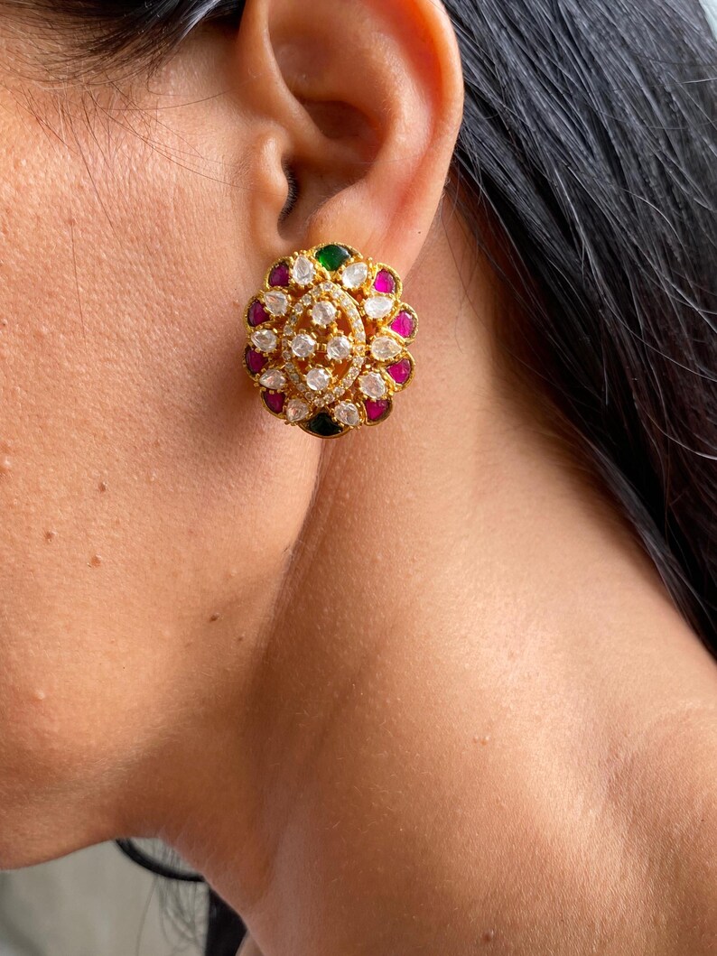 May include: Gold-toned stud earrings with a floral design. The earrings feature a central cluster of clear, sparkling stones surrounded by alternating pink, green, and clear stones. The earrings are set against the skin.