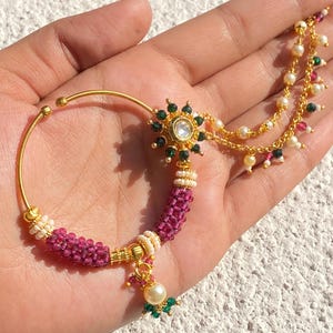 Peut inclure: Un anneau de nez doré avec un design circulaire. L'anneau présente un motif floral central avec une pierre transparente et des accents verts. Une section perlée dans les tons de rose et de blanc est attachée à l'anneau, ainsi qu'une perle et des perles vertes pendantes.