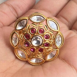 May include: A gold ring featuring a large, round, floral design. The ring is set with clear and pink stones.