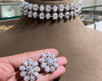 May include: A silver-toned choker necklace and matching earrings set. The choker features two rows of clear, sparkling gemstones in various shapes. The earrings are flower-shaped, with a central rectangular stone surrounded by smaller stones.