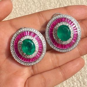 Peut inclure: Une paire de boucles d'oreilles rondes avec une grande pierre ovale verte entourée d'un halo de petites pierres blanches et d'un anneau de pierres roses.
