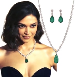 May include: A silver necklace and matching earrings set featuring pear-shaped emerald green gemstones. The necklace has a series of small, clear gemstones leading to a larger emerald pendant. The earrings also have emerald green teardrop gems.