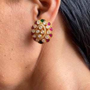 May include: A gold-toned stud earring with a floral design. The center features a cluster of clear, round stones, surrounded by alternating pink, green, and clear stones. The earring is set against the skin.