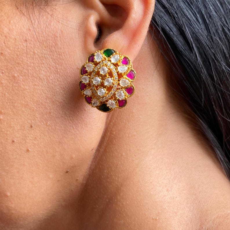 May include: A gold-toned stud earring with a floral design. The center features a cluster of clear, round stones, surrounded by alternating pink, green, and clear stones. The earring is set against the skin.